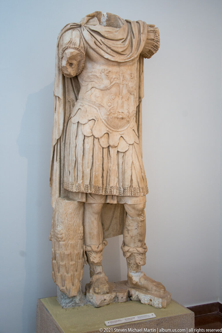 Headless stone male statue wearing a decorated cuirass, short military skirt, and fitted leg coverings.