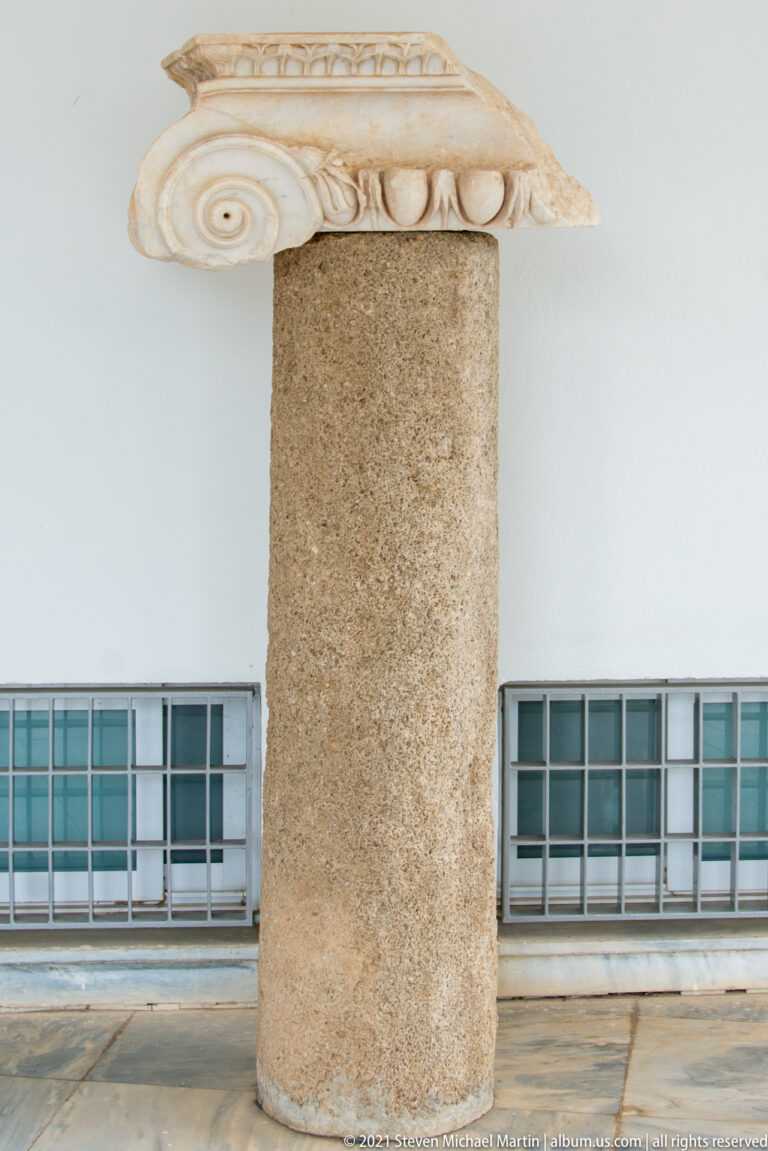 Carved pale stone fragment with spiral volute and egg-shaped ornament displayed on a cylindrical support.