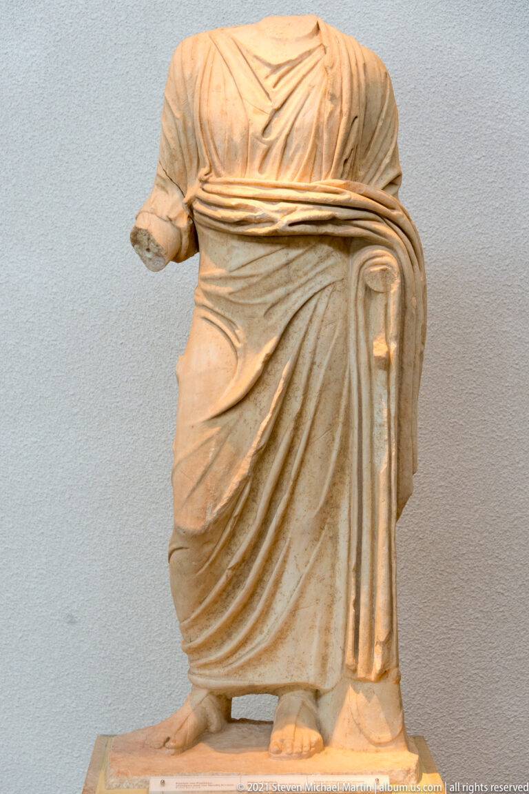 Headless stone woman in a belted long dress and heavy mantle, with deep folds falling beside her legs.