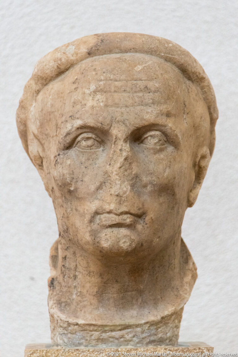 Close frontal view of a weathered stone male portrait head with short hair, large eyes, and straight nose.