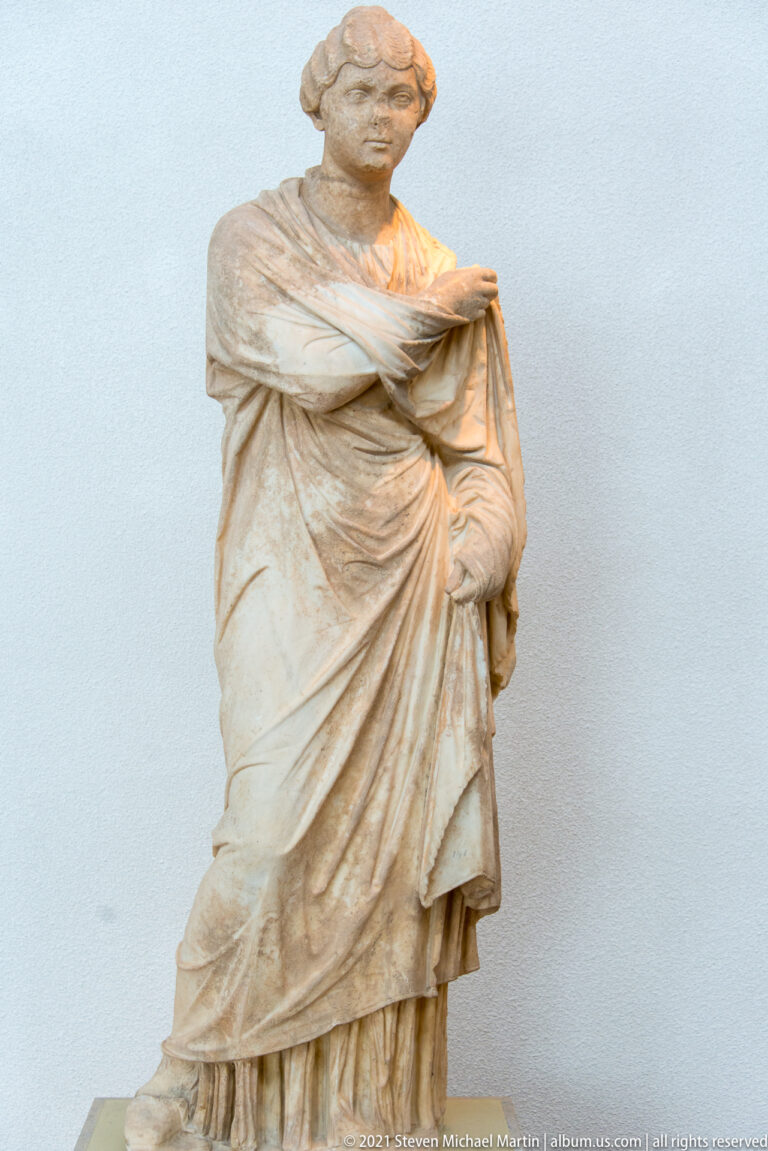 Standing stone woman with surviving head, wrapped mantle, and long layered folds descending to the feet.