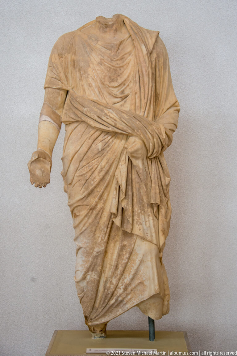 Headless standing stone figure wrapped in a heavy mantle with dense diagonal folds across the torso and waist.