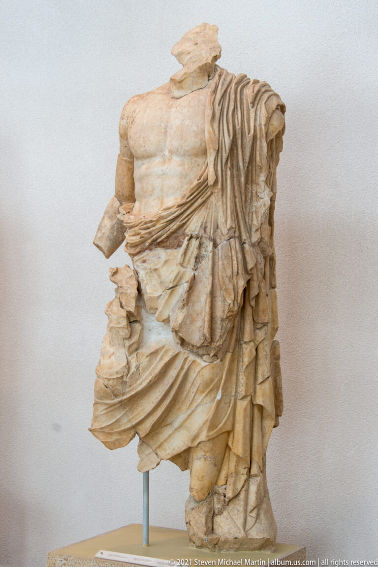 Fragmentary muscular stone male torso with heavy mantle draped over one shoulder and around the lower body.