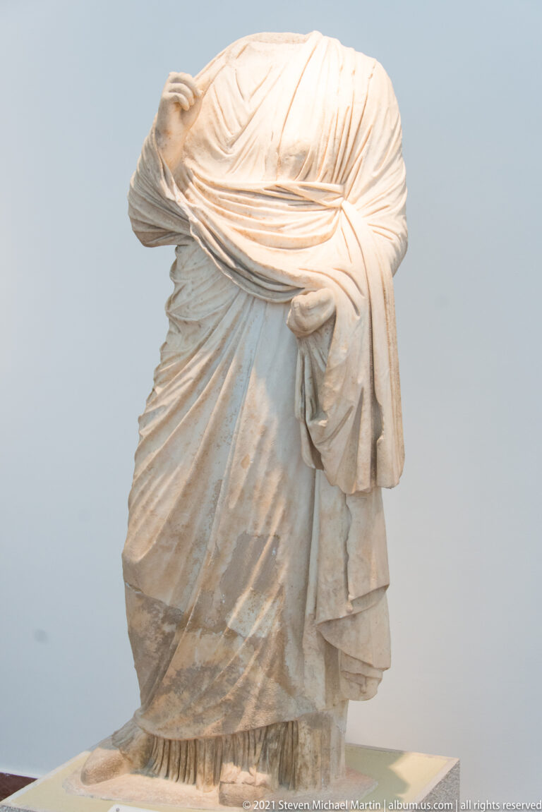 Headless stone woman bending forward, with layered drapery pulled across her back and falling in long folds.