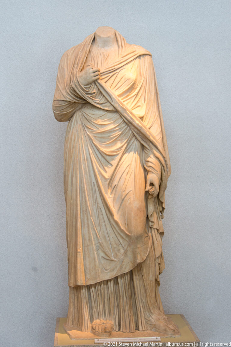 Headless stone woman wrapped in diagonally draped mantle over a pleated long undergarment.