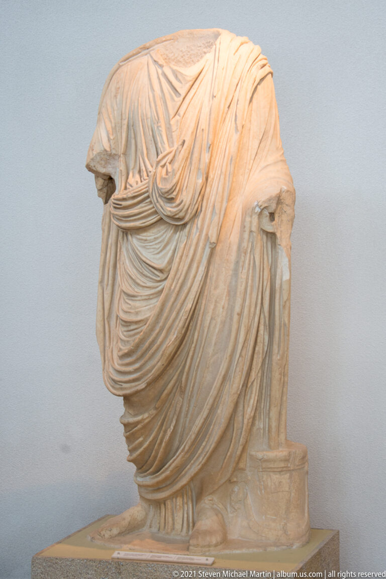 Headless stone woman in deeply looped drapery standing beside a small rectangular support at her right leg.