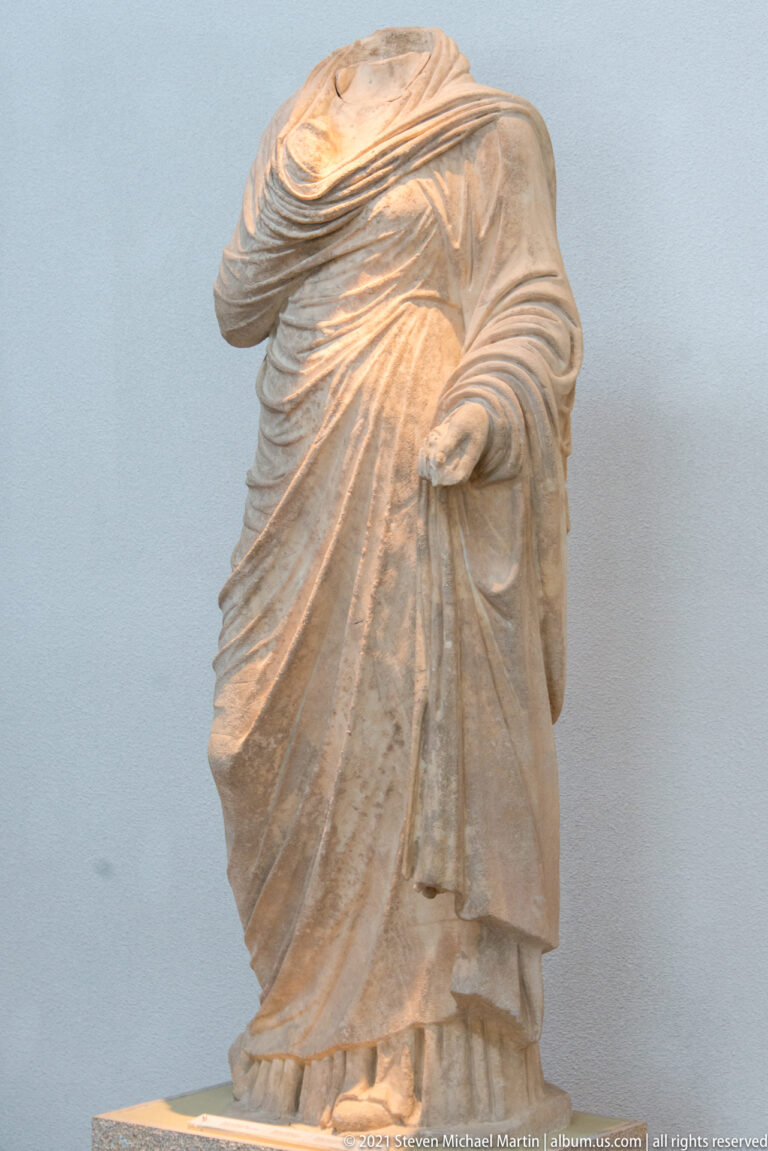 Headless stone woman wrapped in heavy diagonal drapery, with one hand emerging to hold the garment.