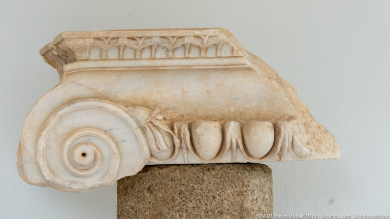 Close view of a carved stone volute beneath a row of egg-shaped architectural ornament.