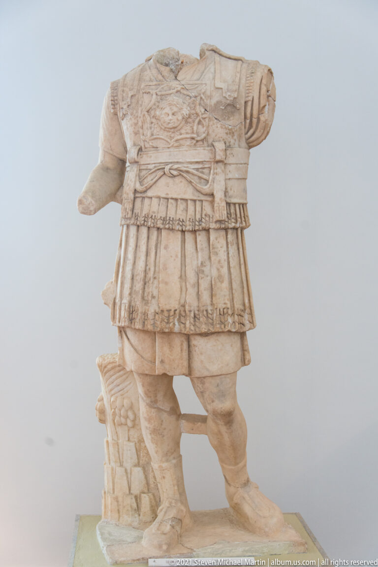 Headless stone man wearing an ornate relief cuirass and layered military skirt, standing beside a sculpted support.