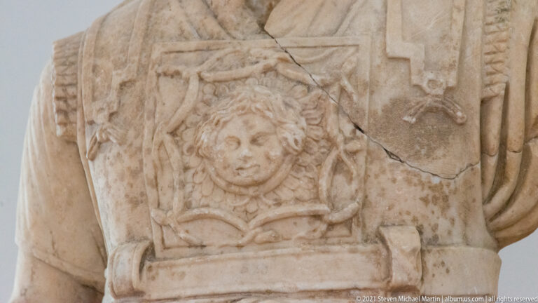 Close stone relief of a Gorgon head with curling hair on the decorated breastplate of an armored statue.