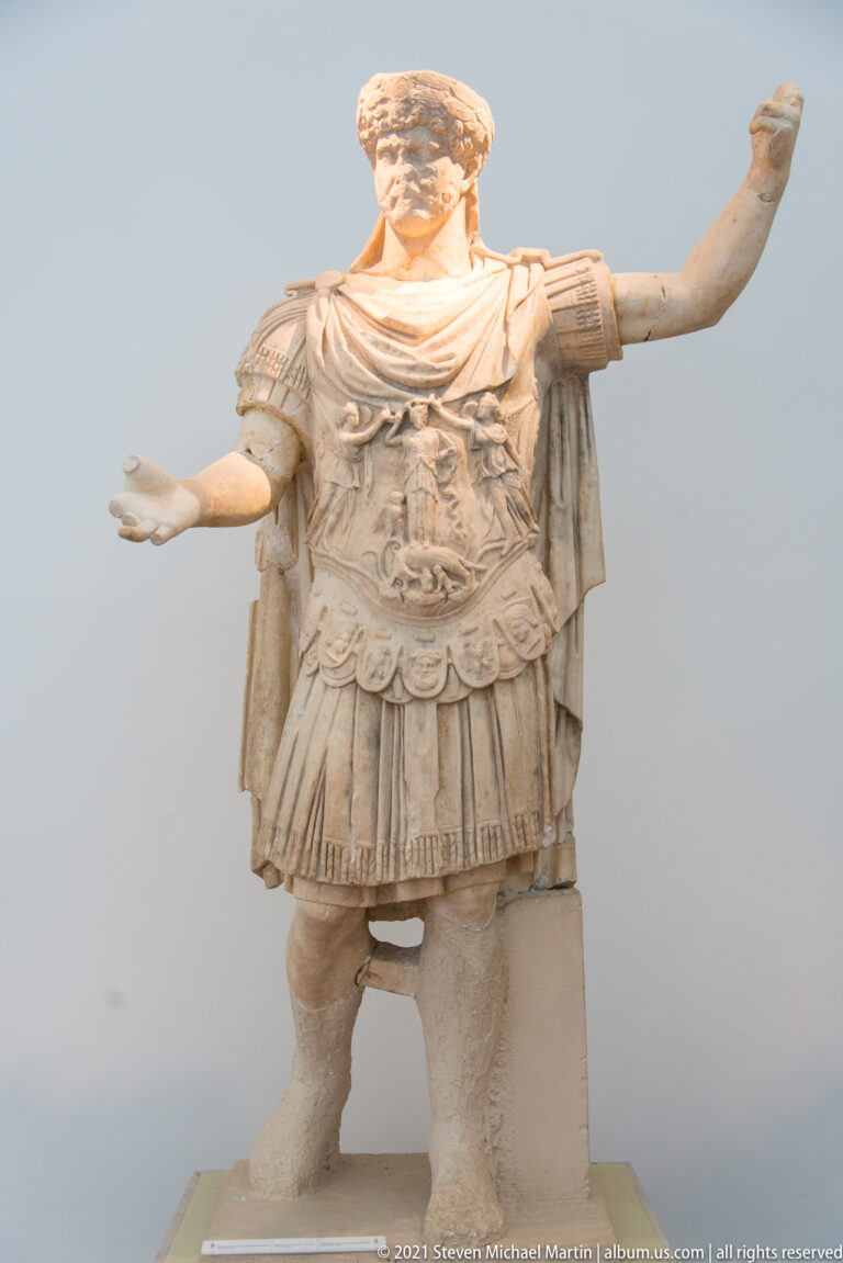 Stone armored man with surviving head, ornate cuirass, cloak, and both arms extended outward.