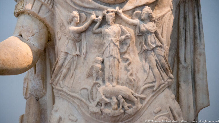 Close detail of a stone cuirass carved with small standing figures, an animal motif, and surrounding symbols.