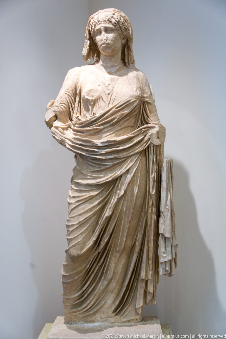 Stone woman with surviving head wrapped in a heavy mantle drawn across her chest and falling in deep folds.