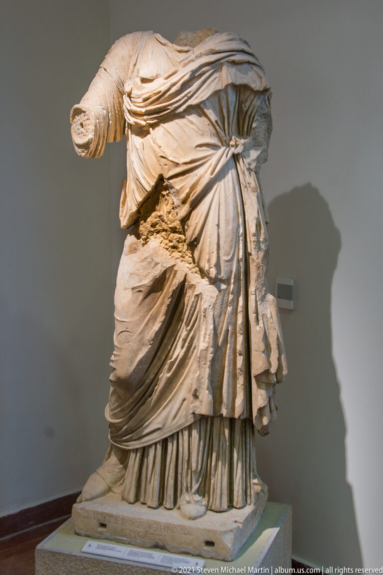 Headless stone woman with layered mantle gathered at one side over a finely pleated long underdress.