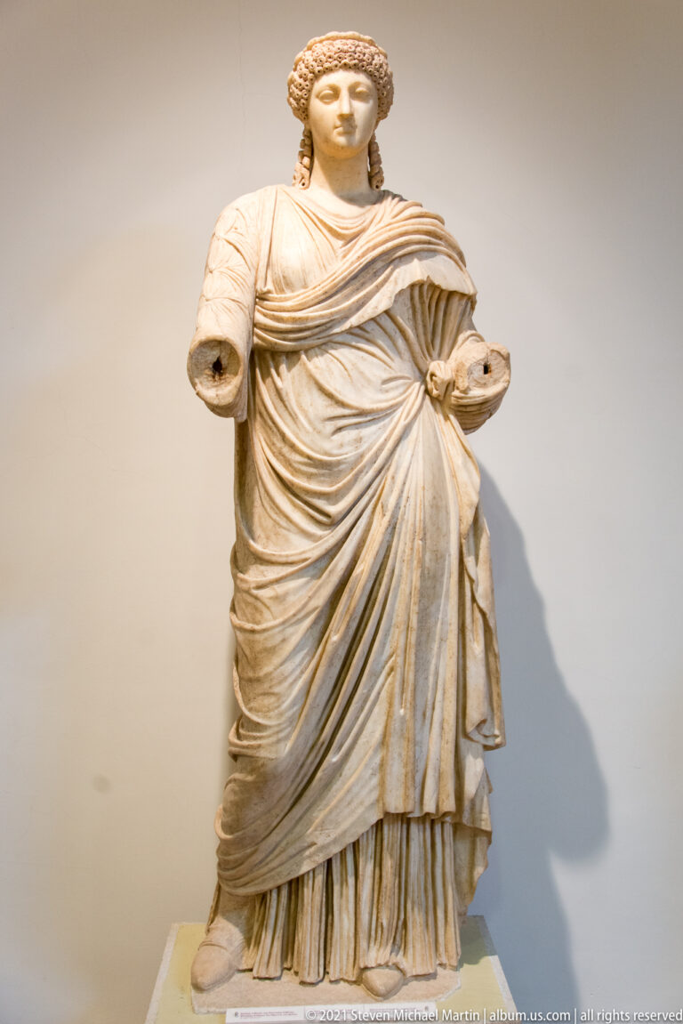 Standing stone woman with surviving head, layered mantle, pleated long dress, and missing forearms.