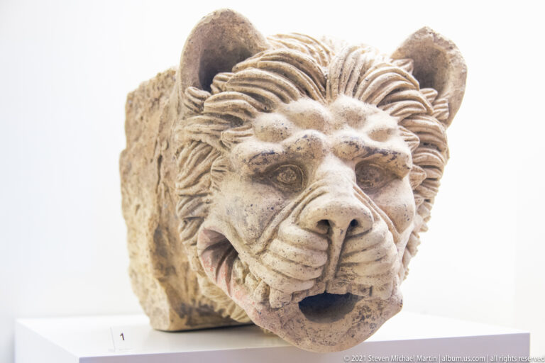 Projecting carved lion head with open drainage mouth, rounded ears, thick mane, and traces of reddish color.