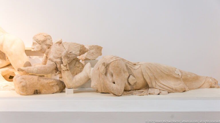 Fragmentary reclining marble figure with draped lower body and scattered adjoining pieces on a museum mount.
