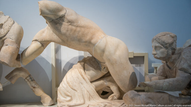 Close museum view of a marble horse's hindquarters above fragmentary human figures in a reconstructed pedimental group.
