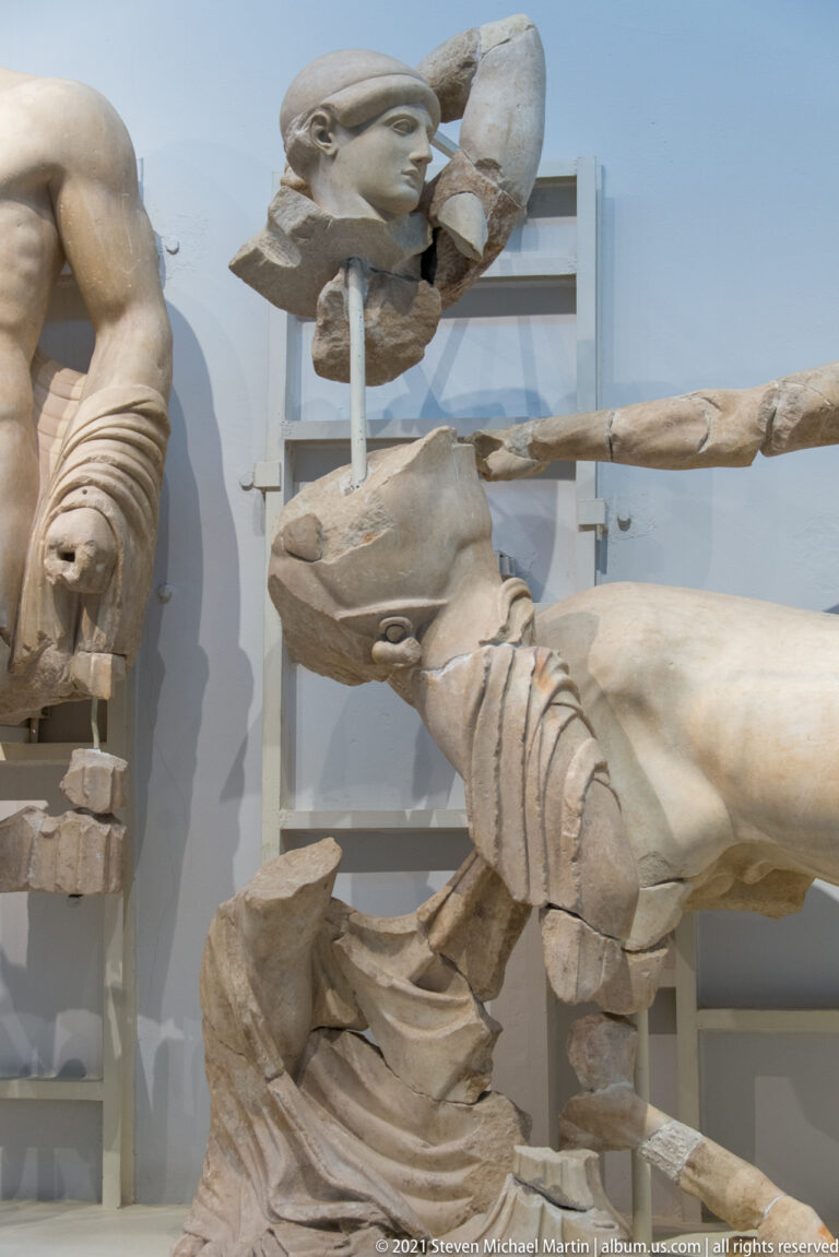 Close museum view of a centaur's horse body with adjacent fragmentary marble figures in a reconstructed pediment.