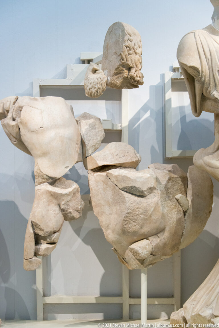 Bearded marble head, torso fragments, and broken limbs mounted separately within a reconstructed Greek temple pediment.
