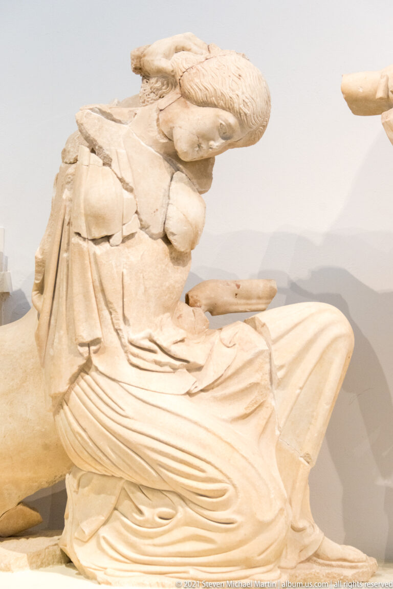 Reclining marble woman in deeply carved drapery with one knee raised, displayed as part of a temple pediment.