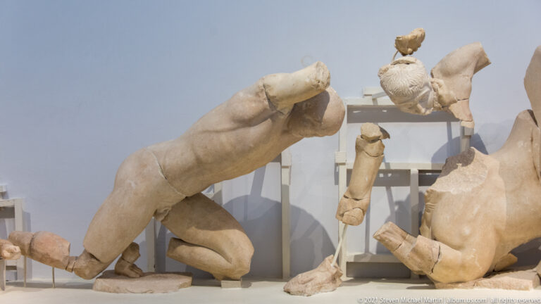 Large fragmentary marble nude figure with detached limbs and nearby sculptural pieces on museum supports.