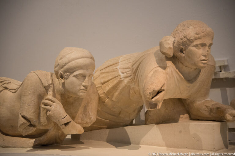 Two reclining marble figures, one wearing a close head covering, positioned along the low edge of a pediment.