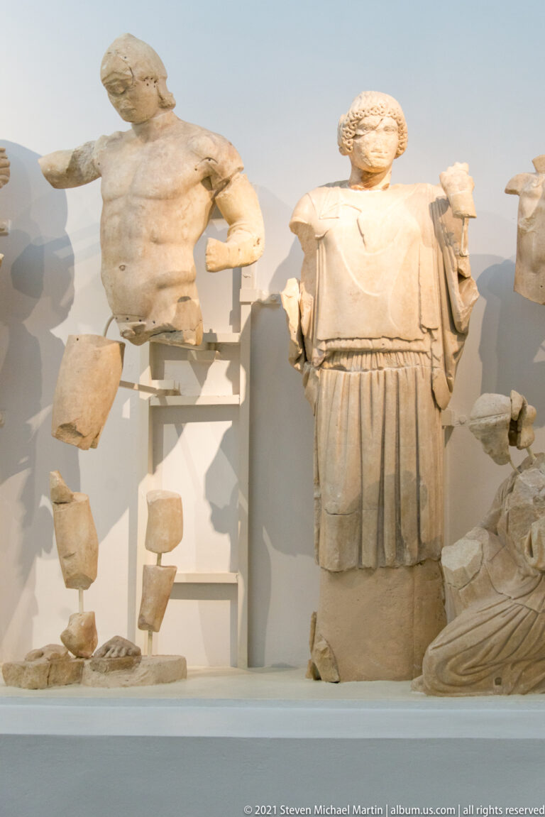 Standing draped marble woman beside a fragmentary nude male torso and detached limbs in a reconstructed pediment.