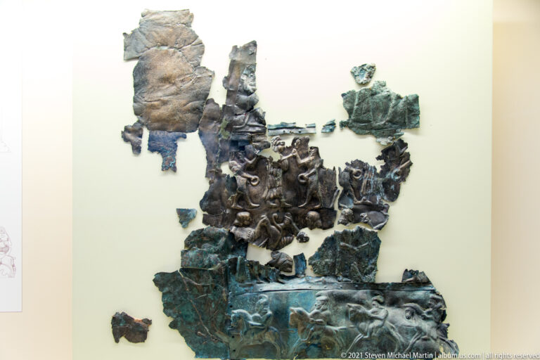 Fragmentary green and dark bronze relief pieces mounted together on a pale museum panel.
