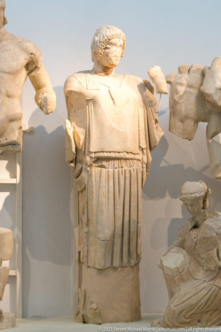 Standing marble woman with surviving head, belted garment, and long vertical folds in a reconstructed pediment.