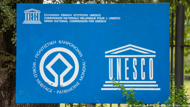 Blue UNESCO World Heritage sign with official emblems and multilingual text at Ancient Olympia.