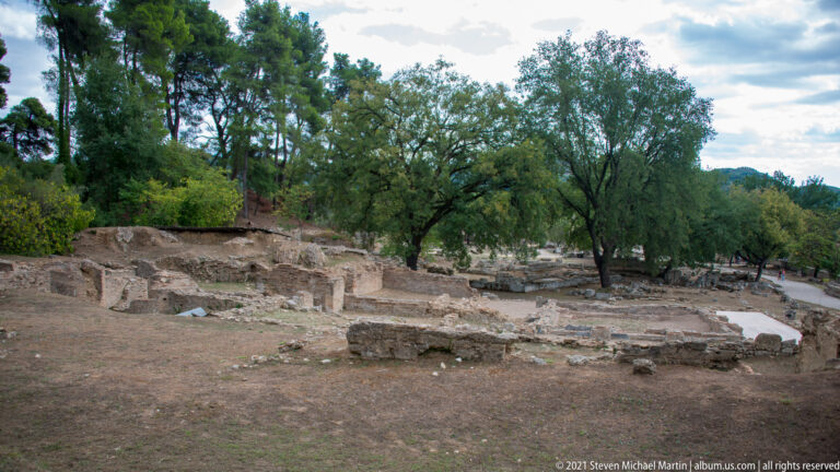 Low stone and brick archaeological foundations extending beneath mature trees at Ancient Olympia.