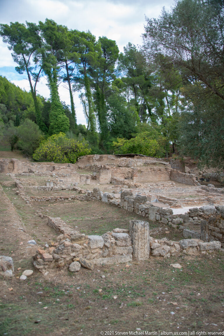 Low stone walls, scattered blocks, and short column fragments beneath trees at Ancient Olympia.
