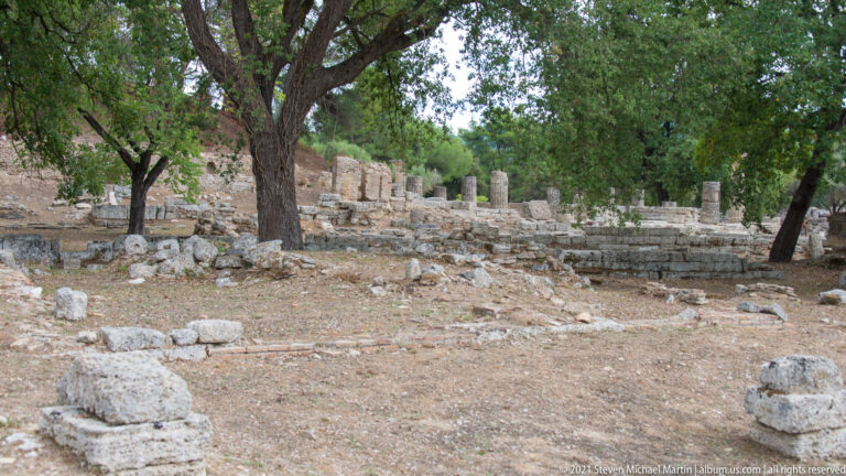 Wooded archaeological area with low stone walls, scattered blocks, and a line of short column stumps.