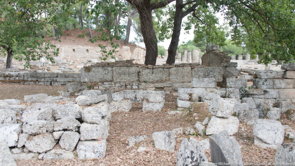 Close view of low stone walls, squared masonry blocks, and scattered fragments beneath trees at Ancient Olympia.