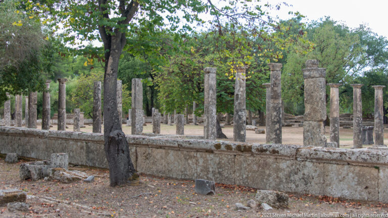 Rows of slender ancient stone columns around an open courtyard amid trees at Ancient Olympia.