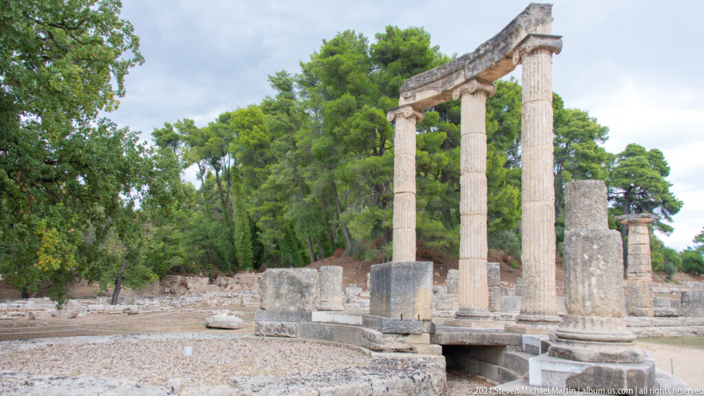 Restored Ionic columns and entablature of the circular Philippeion rising above low foundations and trees at Olympia.
