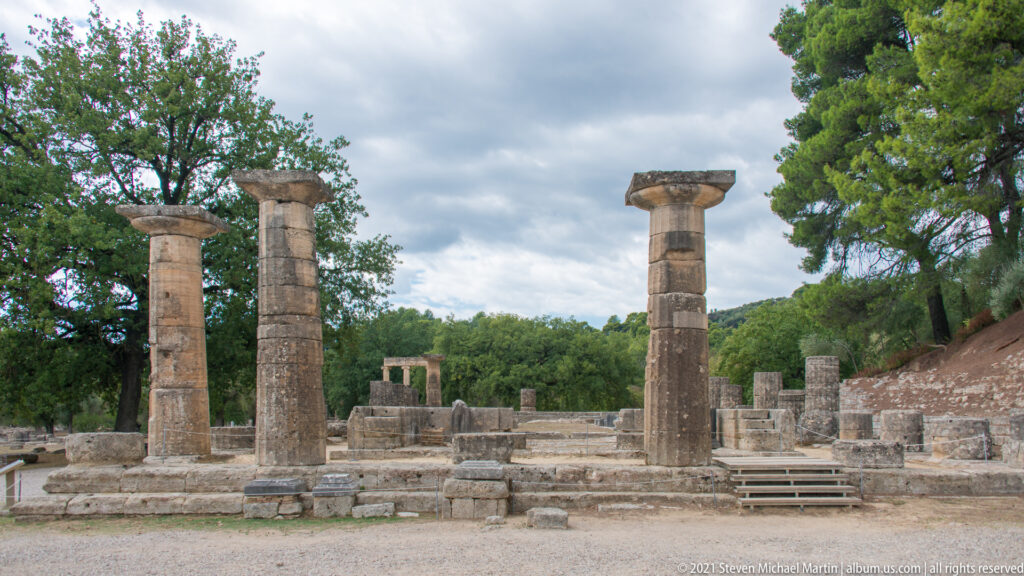 Several tall stone columns among low archaeological walls, platforms, and scattered blocks under a cloudy sky.