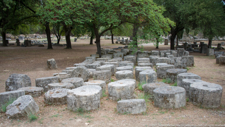Rows of weathered stone column drums and rectangular blocks arranged on the ground beneath trees.