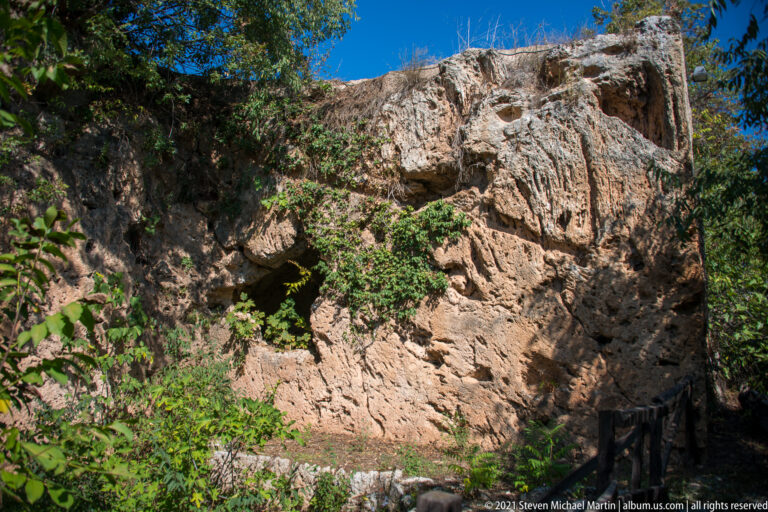 Sunlit rugged rock face with deep cavities, fissures, ledges, and climbing vegetation at the Mieza archaeological site.
