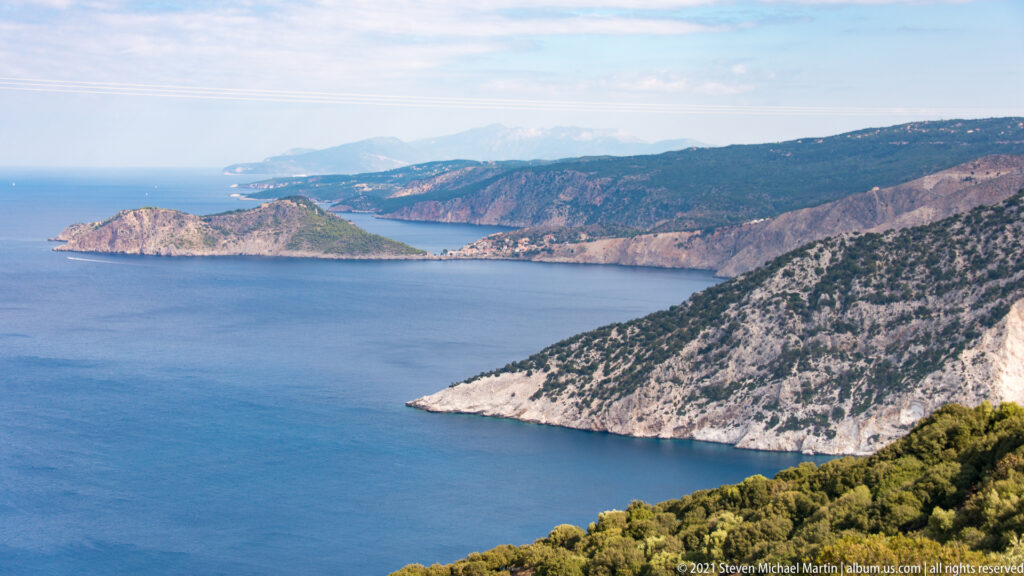 Elevated view across Assos bay toward a pine-covered peninsula, pale limestone cliffs, distant headlands, and calm blue sea.