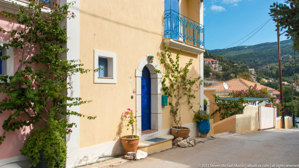 Yellow stucco house with blue arched door, blue balcony, potted plants, and flowering vines on a narrow Assos lane.