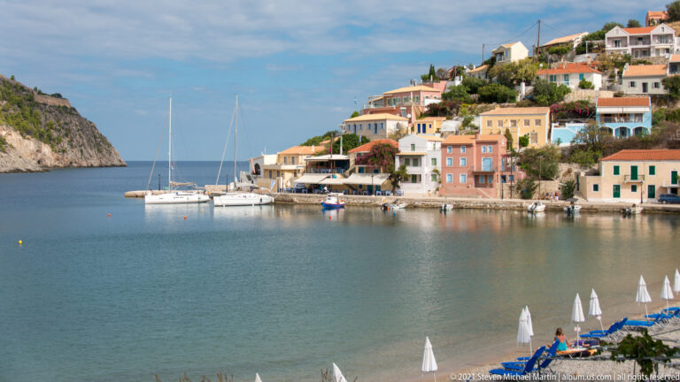 Anchored sailboat in Assos harbor facing pastel waterfront houses, with folded umbrellas and blue lounge chairs in the foreground.