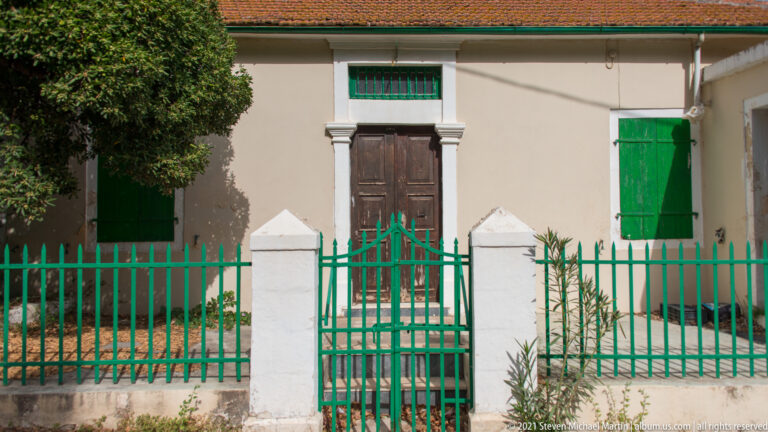 Stucco house with central dark wooden doors, green shutters, red-tiled roof, and green iron fence in Assos.