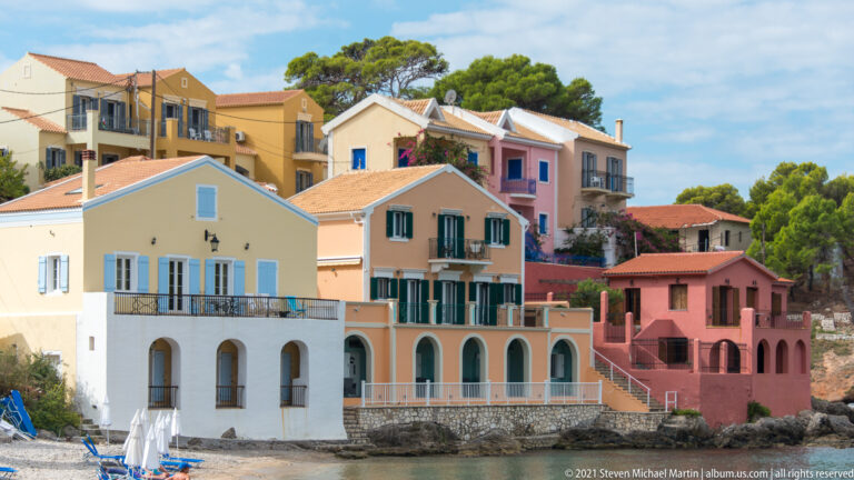 Yellow, peach, and rose waterfront residences with arches, balconies, shutters, and red-tiled roofs beside the sea in Assos.