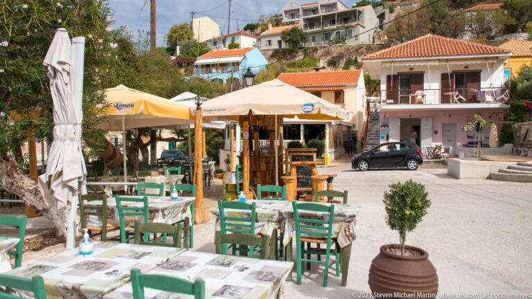 Outdoor dining area with green chairs, set tables, cream umbrellas, trees, and pastel buildings in Assos.