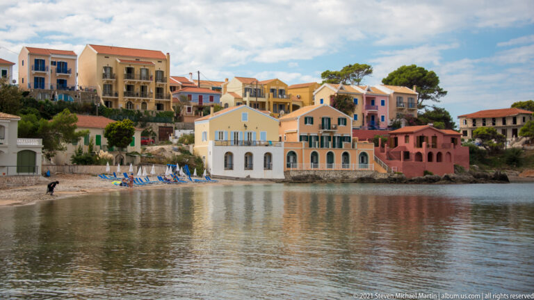 Pastel waterfront residences reflected in calm Assos water beside a narrow beach lined with blue lounge chairs.