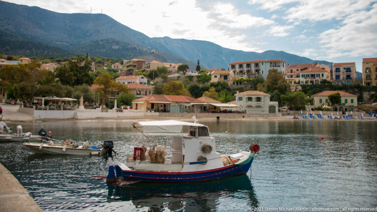 Small blue-and-white open motorboat in Assos harbor, with waterfront buildings and hillside houses in the background.