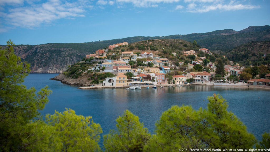Elevated view of Assos village on a small waterfront promontory, framed by pine trees and backed by forested mountains.
