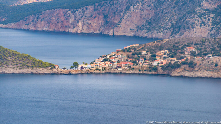 Distant view of Assos village clustered along a sheltered cove beneath steep rocky mountains.